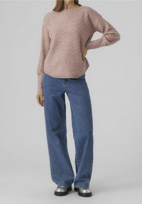 Mujer con un suéter texturado rosa, jeans azul holgados y zapatos de suela gruesa plateados, de pie con una mano en la cadera.