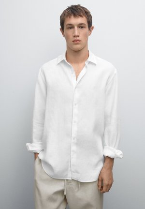 Weißes Leinenhemd mit langen Ärmeln, Knopfleiste, spread collar und aufgeschlagenen Manschetten, kombiniert mit hellbeigen Hosen.