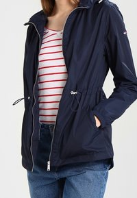 Giacca a vento blu navy con zip frontale, cordino regolabile in vita e due tasche laterali. Presenta un cappuccio e include dettagli in metallo argentato.