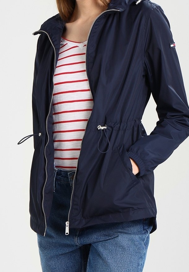 Giacca a vento blu navy con zip frontale, cordino regolabile in vita e due tasche laterali. Presenta un cappuccio e include dettagli in metallo argentato.