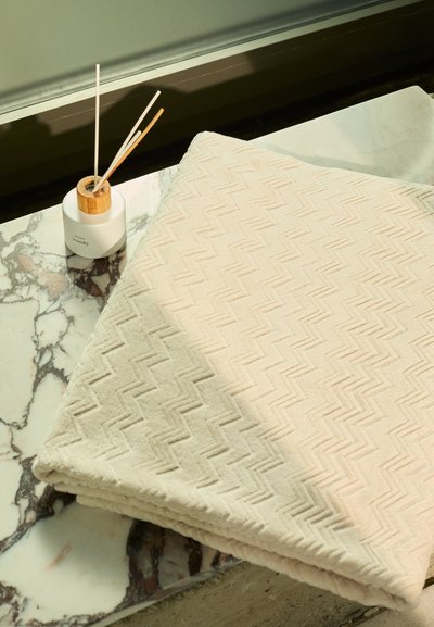 Beige színű törölköző, zigzag mintával, puha anyagból, egy márványfelületen elhelyezve, mellette egy henger alakú tároló edénnyel, amelynek fából készült díszítése van.