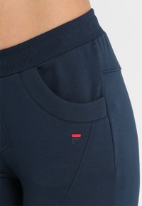 Mörkblåa sweatpants med ribbad midjeresår, sidoficka och en liten logotyp i rött och svart. Slät, stretchig tyg.