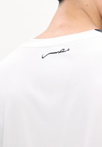Gros plan du cou et du haut du dos portant une chemise blanche avec une écriture arabe noire près du col.