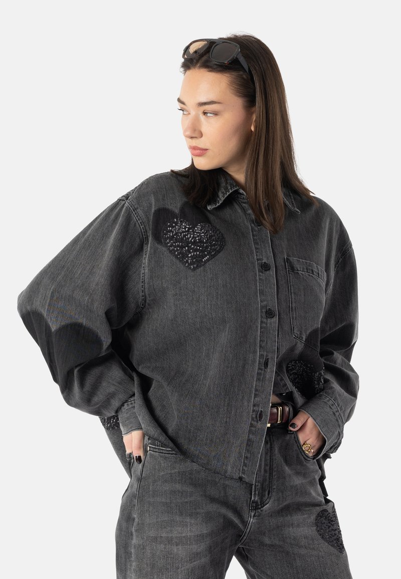 Camicia oversize in denim nero con grandi accenti a forma di cuore in paillettes nere, chiusura con bottoni e tasca applicata. Abbinata a pantaloni in denim nero coordinati.