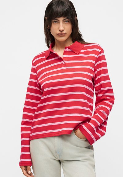 Νεαρή γυναίκα με σκούρα μαλλιά που φοράει κόκκινο μπλούζα πόλο μακρυμάνικη με λευκές οριζόντιες ρίγες, ανοιχτόχρωμο παντελόνι, χέρι στην τσέπη.