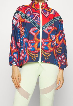 Vrouw draagt een kleurrijke Adidas jas met rits en bloemmotieven, gecombineerd met lichtgekleurde leggings met doorschijnende meshpanelen.