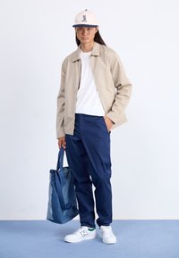 Beige bomber dzseki, fehér póló, sötétkék nadrág, fehér edzőcipő zöld részletekkel. Kék farmer vászontáska van nála, világoskék felületen áll.
