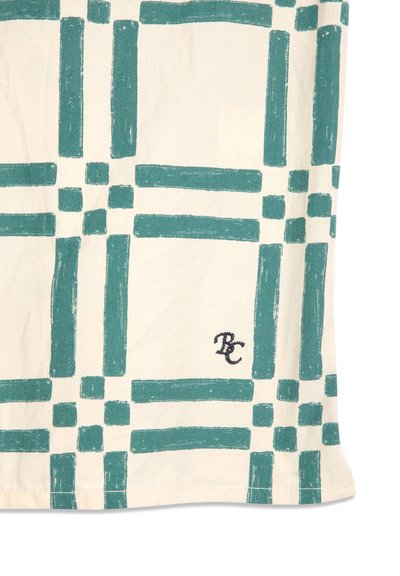 Tissu écru avec motif à carreaux verts irréguliers et petites initiales "RC" noires brodées près du bord inférieur.