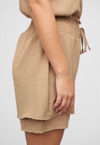 Robe en tissu beige froissé avec une taille ajustée et un cordon de serrage. Texture douce et coupe ample se terminant par un ourlet en couches.