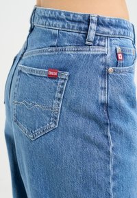 Hellblaue Jeans mit hoher Taille, versehen mit einem roten Logo-Aufnäher auf der hinteren Tasche und sichtbaren Nähdetails an den Taschen.