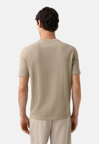 Homme avec des cheveux courts et bouclés portant un pull beige à manches courtes et un pantalon beige clair, se tenant de dos devant un fond neutre.