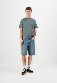 Grå t-shirt med blå grafik på venstre bryst, parret med blå denimshorts og sorte sneakers. Har hvide sokker og et afslappet design.