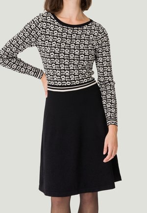 Femme portant un haut à manches longues noir et blanc à motifs avec le texte "NOTION" et une jupe noire unie jusqu'aux genoux.