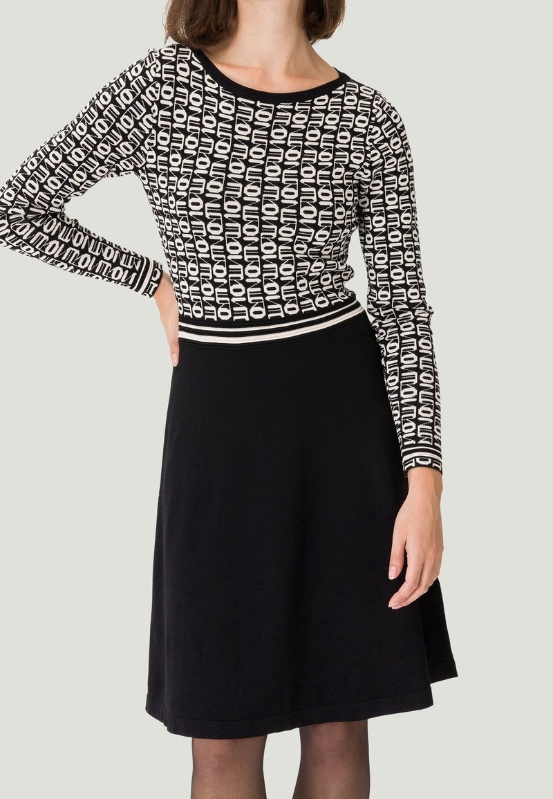 Femme portant un haut à manches longues noir et blanc à motifs avec le texte "NOTION" et une jupe noire unie jusqu'aux genoux.