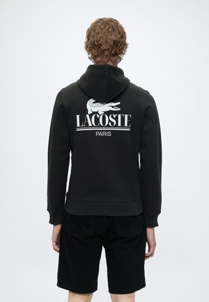 Personne aux cheveux bouclés portant un sweat à capuche noir Lacoste Paris et un short noir, vue de dos sur un fond uni.
