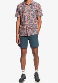 Koszula z krótkim rękawem w kolorach koralowym i granatowym z geometrycznym wzorem, zestawiona z ciemnymi szortami. Do tego paskowane skarpetki i czarne buty sportowe.