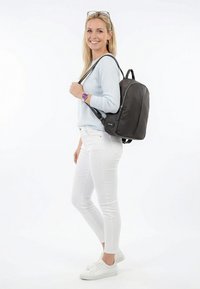 Naine seljas hele sinine kampsun, valged püksid ja tossud, naeratades, kandes ühel õlal musta seljakotti, taust on valge ja lihtne.