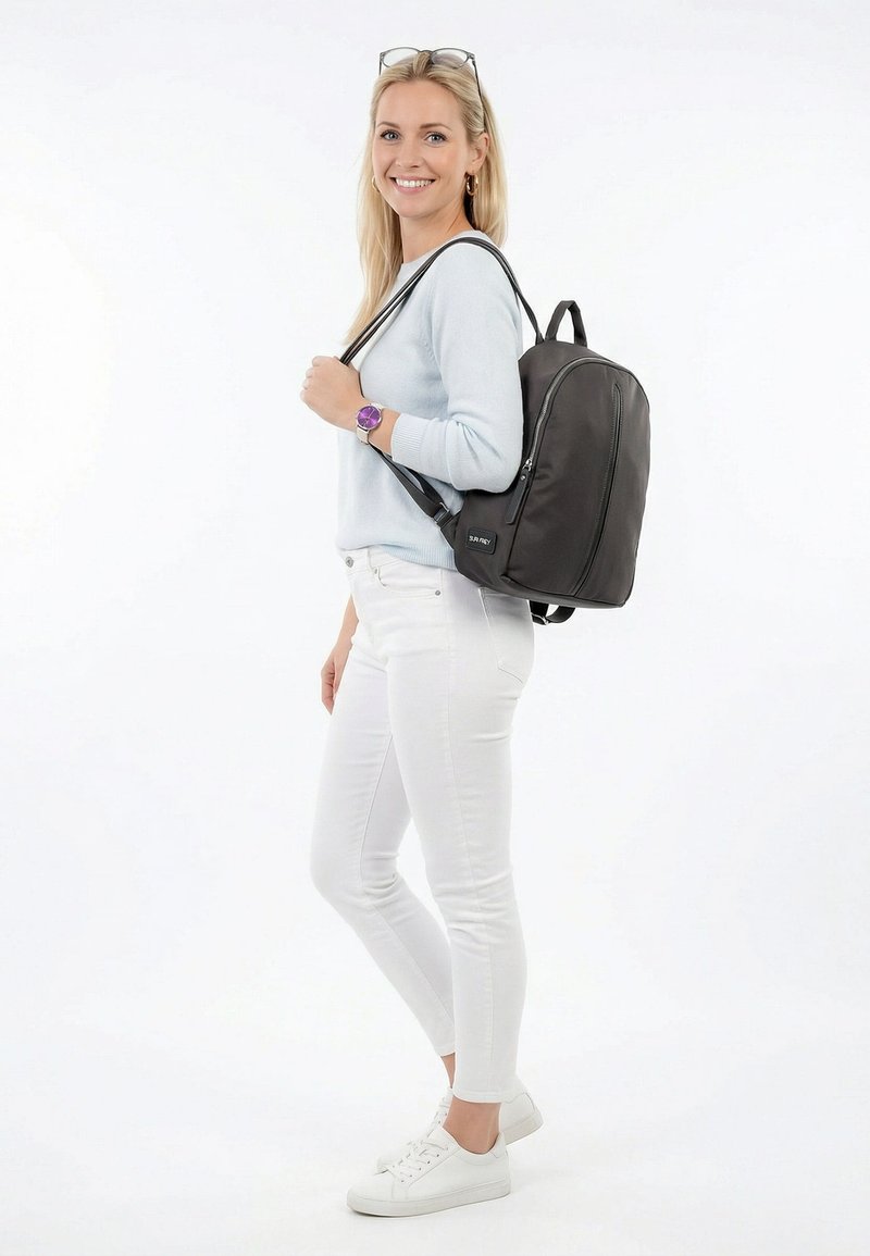 Naine seljas hele sinine kampsun, valged püksid ja tossud, naeratades, kandes ühel õlal musta seljakotti, taust on valge ja lihtne.