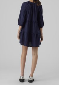 Kvinna som står, klädd i en lös mörkblå lager-på-lager klänning med trekvartslång ärm och silverfärgade klumpiga skor.