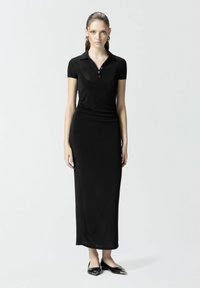 Crna maksi haljina s polo vrhom kratkih rukava, izrađena od glatkog materijala. Ima prilagođen siluet i elegantne ravne cipele s špicastim vrhom.