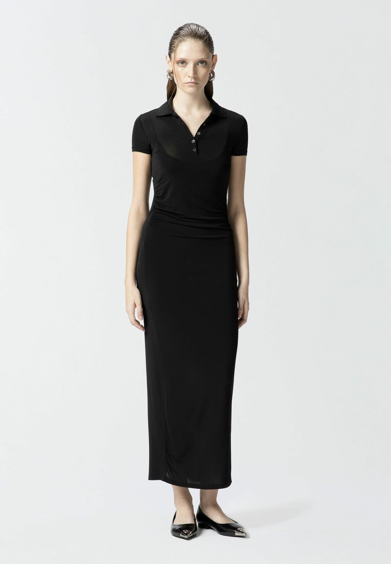 Crna maksi haljina s polo vrhom kratkih rukava, izrađena od glatkog materijala. Ima prilagođen siluet i elegantne ravne cipele s špicastim vrhom.