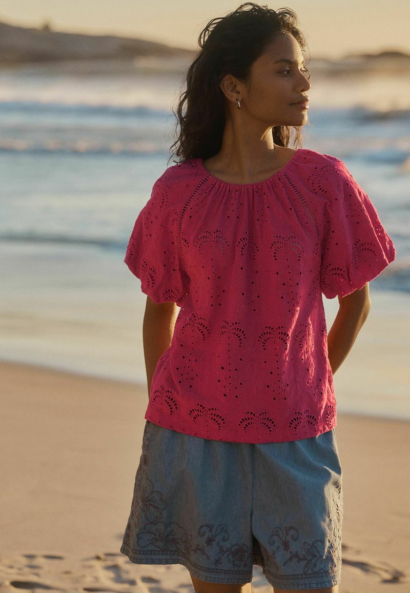 Femme aux cheveux bouclés foncés portant un chemisier rose ajouré et un short gris brodé de fleurs, se tenant sur une plage au coucher du soleil.