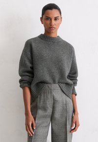 Grauer Pullover mit geribbeltem Kragen, überschnittenen Schultern und strukturierter Oberfläche. Kombiniert mit grauen karierter Hose mit vorderen Falten.