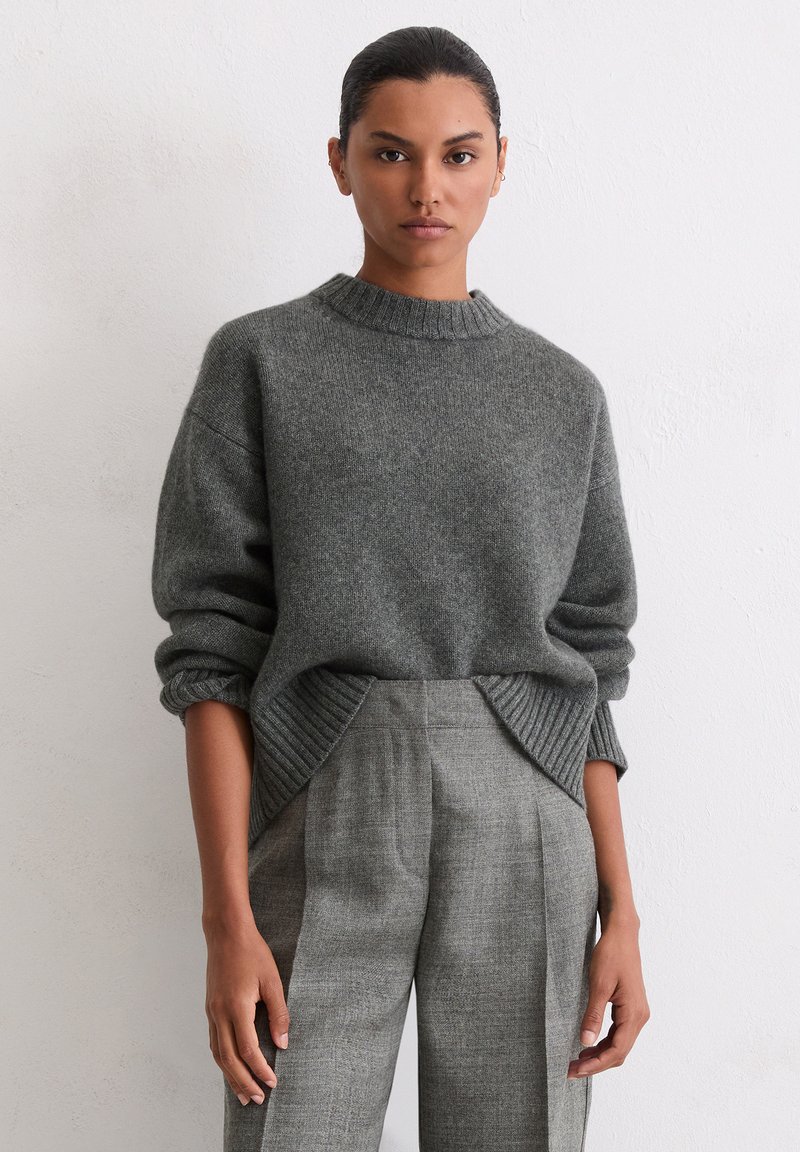 Grauer Pullover mit geribbeltem Kragen, überschnittenen Schultern und strukturierter Oberfläche. Kombiniert mit grauen karierter Hose mit vorderen Falten.