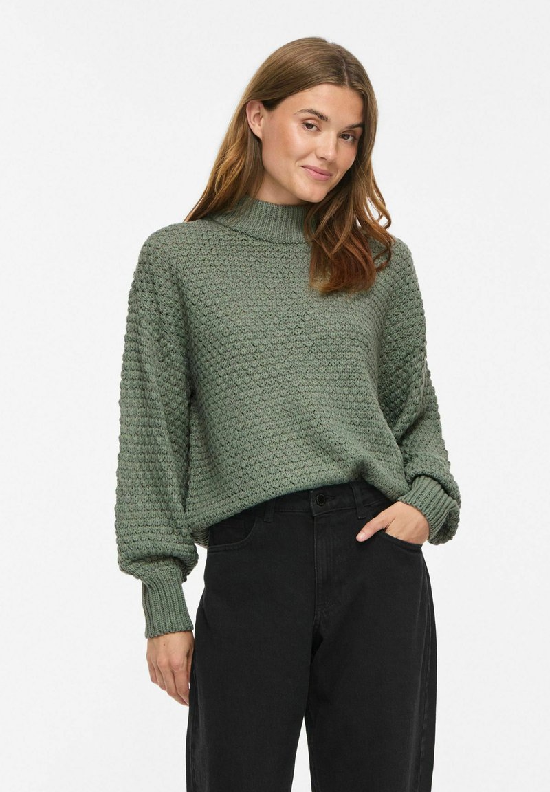 Femme aux cheveux châtain clair portant un pull en maille verte texturée et un pantalon noir taille haute, debout avec une main dans la poche.