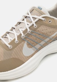 Scarpa da corsa Nike beige e marrone chiaro con suola bianca, lacci bianchi, dettagli in filo blu e tomaia in tessuto a rete con il logo swoosh di Nike.