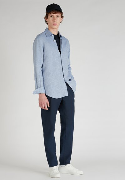 Chemise boutonnée bleu clair avec des manches retroussées, associée à un pantalon bleu marine et des baskets blanches. La chemise a un aspect en lin texturé.