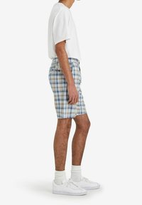 Men's plaid shorts in light blue, white, and yellow. Made of woven fabric with a button closure and side pockets. Paired with a white t-shirt.