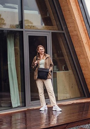 Brown fleece jacket, light green top, beige pants, white sneakers, and black shoulder bag. Wooden building and glass doors in background.
