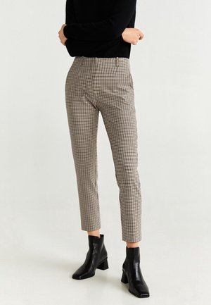 Osoba ubrana w czarną bluzkę z długim rękawem, kraciaste spodnie do połowy łydki i czarne botki, stojąca na jednolitym jasnym tle.