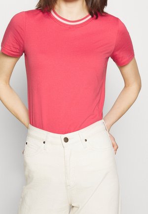 Personne portant un t-shirt rose à manches courtes avec un col rayé blanc, vêtue de jeans taille haute blancs cassés avec les mains dans les poches arrière.