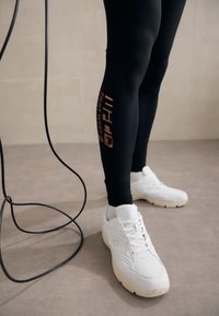 Leggings noirs avec texte et logo orange sur le côté, associés à des chaussures de sport blanches avec une tige texturée et une semelle contrastante.