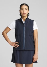 Námornícky vest s vysokým golierom, plným zipsom so modrým akcentom a vrstveným dizajnom, nosený cez bielu košeľu a sukňu.