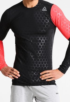 Mann trägt ein schwarz-rotes langärmeliges Reebok-Sportshirt mit sechseckigem Muster und hellgraue Reebok-Shorts, Hände in der Hüfte.