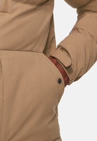 Béžová pufovací bunda s texturovanou tkaninou, která má detail se zapínáním na patky na kapse a nastavitelné manžety se suchým zipem.