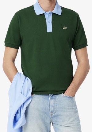 Mand iført mørkegrøn polo med lyseblå krave, holder en matchende lyseblå skjorte og lyseblå jeans.