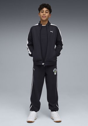 Ragazzo in piedi con le mani nelle tasche che indossa una tuta Puma nera con strisce bianche e sneakers bianche su sfondo grigio.