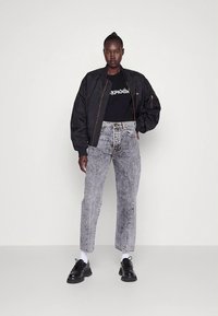 Kvinde står op ad en ensfarvet væg iført sort bomberjakke, sort t-shirt med grafik, højtaljede acid-wash jeans, hvide sokker og sorte platformsko.