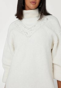 Maglione bianco oversize con colletto a dolce vita, texture a costine e un motivo di pizzo sullo scollo. Materiale in maglia soffice, maniche a tre quarti.