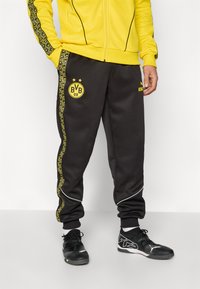 Person iført sorte BVB 09 træningsbukser med gule mønstrede side-striber, parret med sorte sportssko og en gul jakke.
