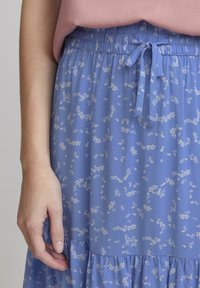 Närmre bild av en person som bär en blå blommig kjol med ett knutet midjeband och en rosa topp, med handen avslappnad bredvid kjolen.