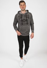 Grå huvtröja med svart grafiskt tryck, långa ärmar och känguruficka, tillsammans med svarta tajta byxor och vita sneakers.