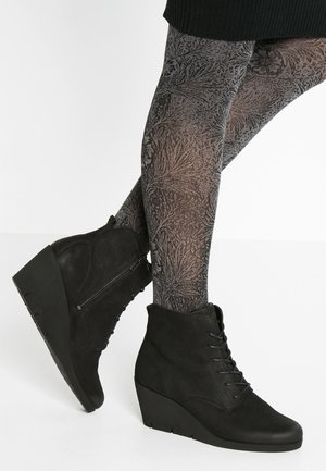 Bottines compensées noires avec fermeture éclair latérale portées avec des collants gris foncé à motif sur des jambes croisées.