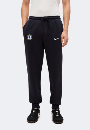 Mężczyzna noszący czarne spodnie dresowe Chelsea FC Nike z elastycznymi ściągaczami, sznurkiem w talii i czarne sneakersy Nike z białym logo.