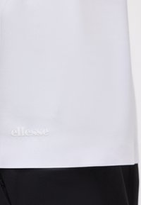 Λευκό ύφασμα με μικρό ανάγλυφο λογότυπο "ellesse" κοντά στο κάτω μέρος πάνω από μαύρο ύφασμα.