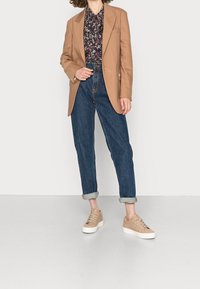Femme portant un blazer camel, un blouse à fleurs, un jean bleu nuit retroussé et des baskets beiges, se tenant devant un fond blanc sobre.